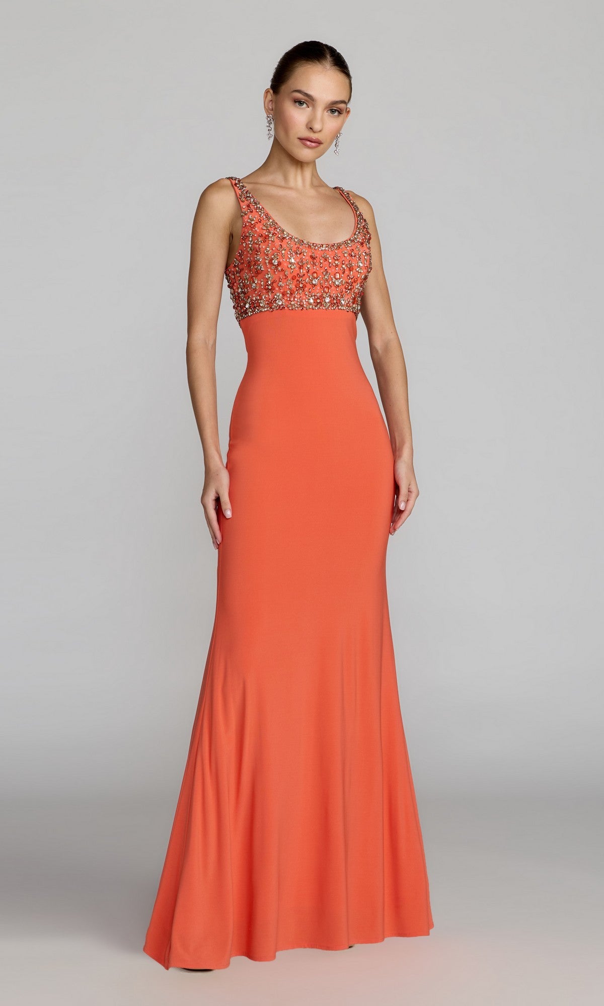 A woman models the Mac Duggal 2253, a long formal dress in coral featuring a sleeveless, floor-length design with a fitted, crystal-embellished bodice and flared skirt, standing against a plain gray background—a sophisticated choice for evening events.